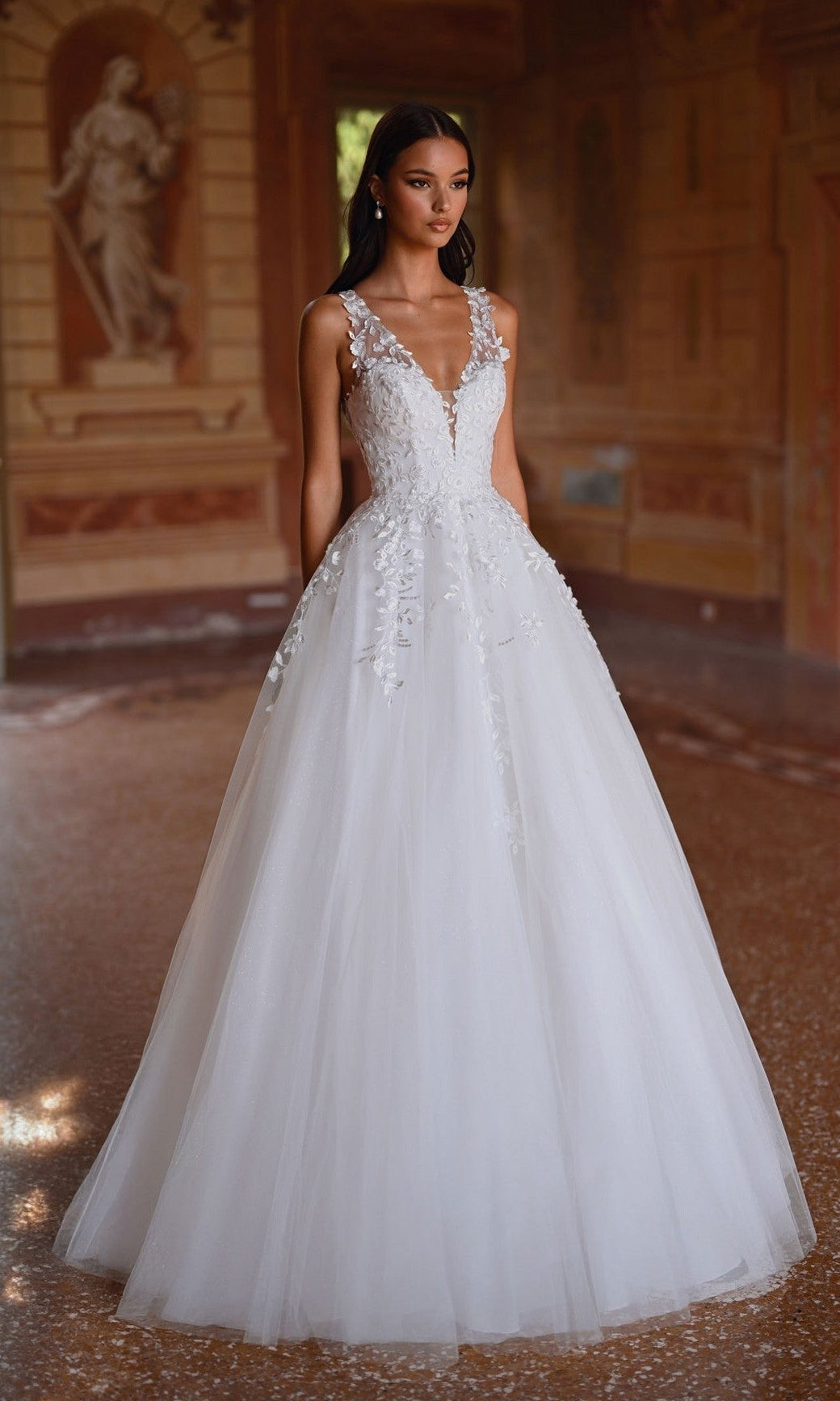 A woman in the Alyce 7128 long ivory dress stands indoors on a polished floor, with classical artwork in the background.